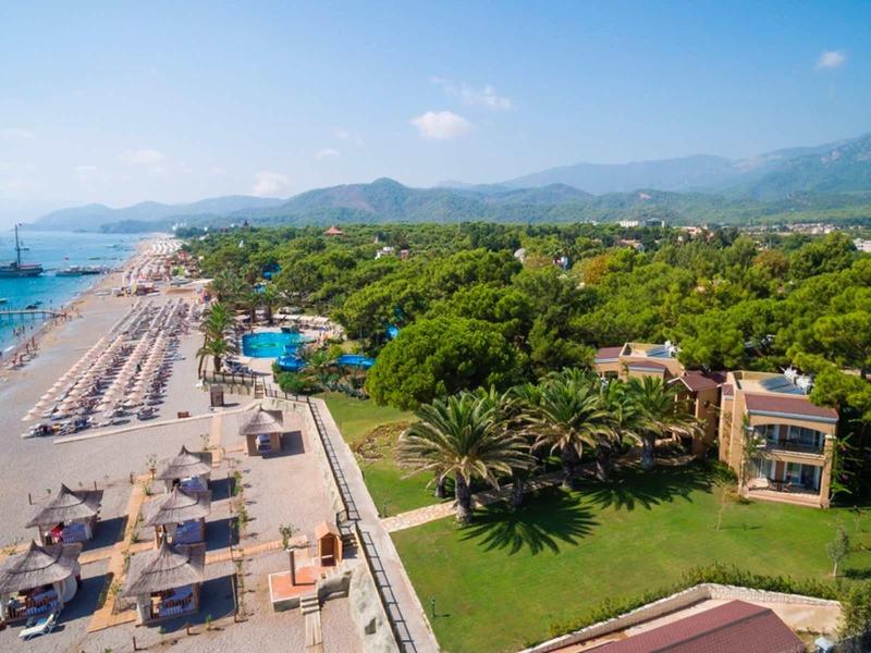 Přímořský hotel s lehátky a zelenými zahradami vedle čistého modrého moře a hor v pozadí.