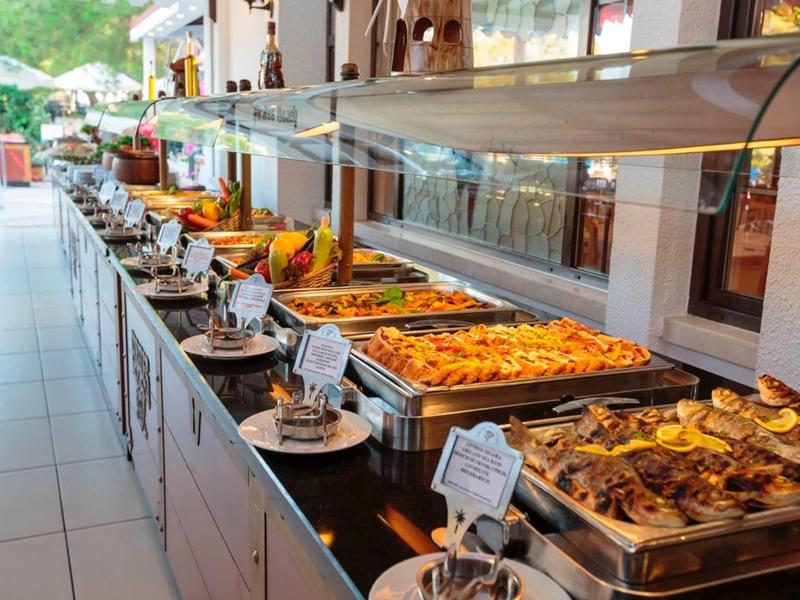 Bufet s různými teplými jídly v nerezových nádobách, podávaný v světlé místnosti.