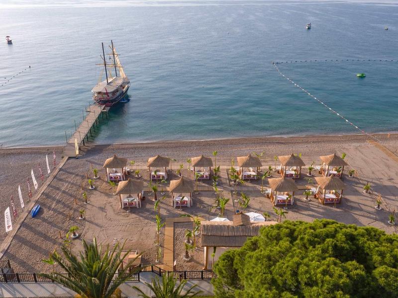 Pohled na pláž s slunečníky, lehátky a molem s plachetnicí na klidném moři.