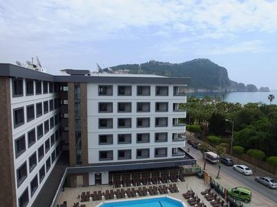 Modern hotelgebouw met meerdere verdiepingen, uitzicht op zee, bomen en heuvels op de achtergrond.