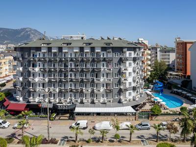 Grand bâtiment d'hôtel avec balcons, piscine et montagnes en arrière-plan par une journée ensoleillée.