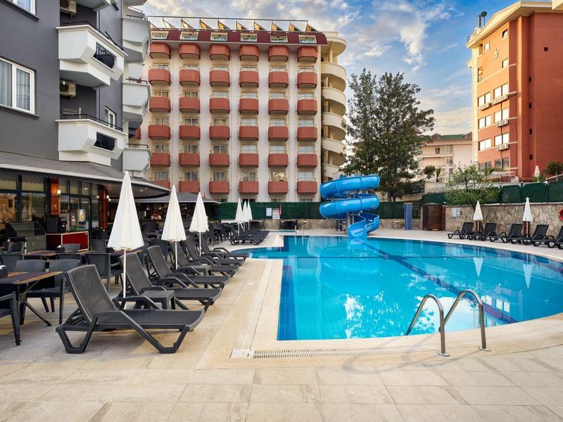 Piscine d'hôtel moderne avec toboggan bleu, chaises longues et parasols près de grands bâtiments.