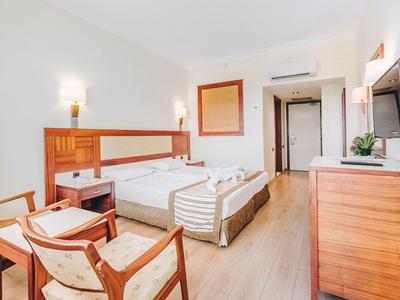 Chambre d'hôtel lumineuse avec lit double, meubles en bois, fauteuils et télévision à écran plat.