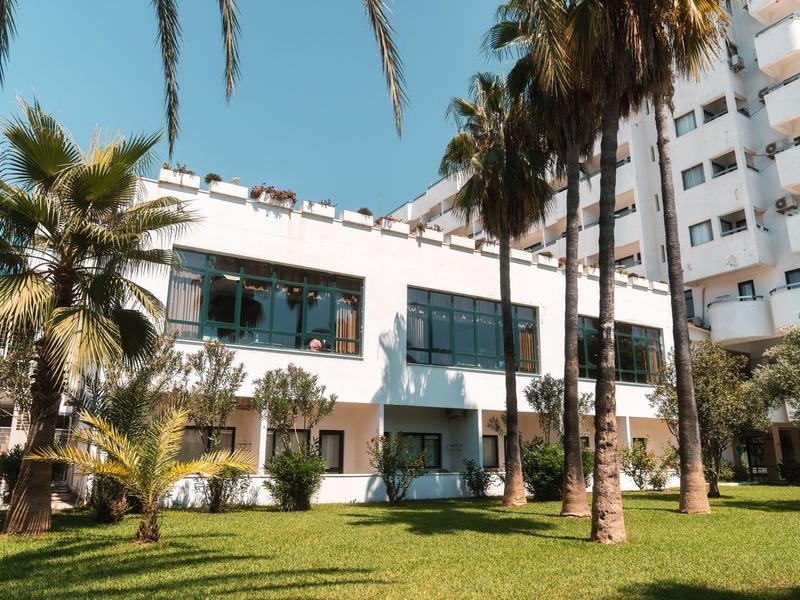 Bâtiment d'hôtel avec grandes fenêtres entouré de palmiers et pelouse verte sous ciel clair.