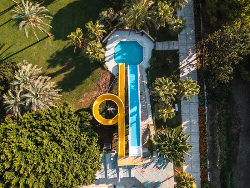 Vue aérienne d'un toboggan bleu et jaune dans un jardin luxuriant avec des palmiers.