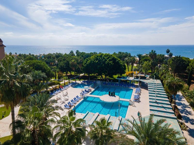 Piscine extérieure d'hôtel entourée de transats, palmiers et vue sur la mer sous un ciel partiellement nuageux