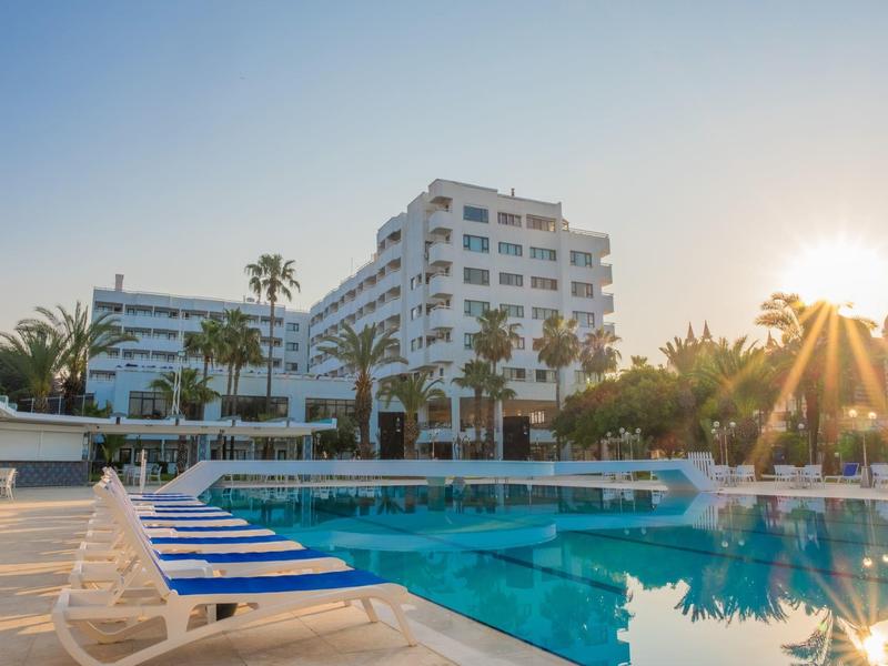 Hôtel moderne avec grande piscine, palmiers et chaises longues sous un ciel clair au coucher du soleil.