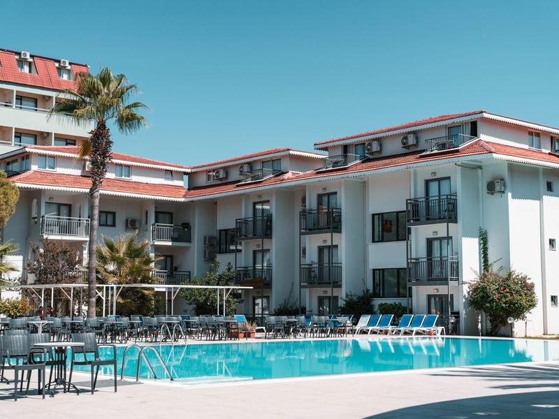 Hotel met buitenzwembad, palmbomen en blauwe lucht in een zonnige omgeving.