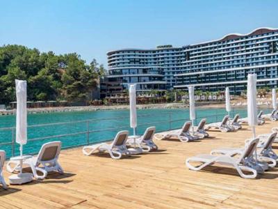 Sonnige Holzterrasse mit Liegestühlen am klaren Wasser vor modernem Hotel und grünen Bäumen.