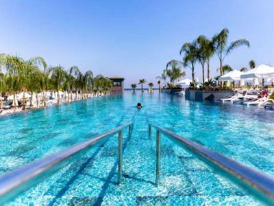Großer, klarer Pool mit Palmen, weißen Liegen und Sonnenschirmen unter blauem Himmel.