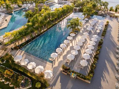 Großer Pool mit weißen Sonnenschirmen und Palmen in einem sonnigen Hotelressort.