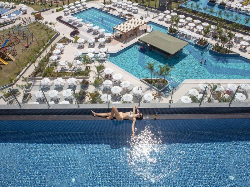 Blick auf ein großes Schwimmbad mit Sonnenschirmen, Liegen und einer Person im Infinity-Pool.