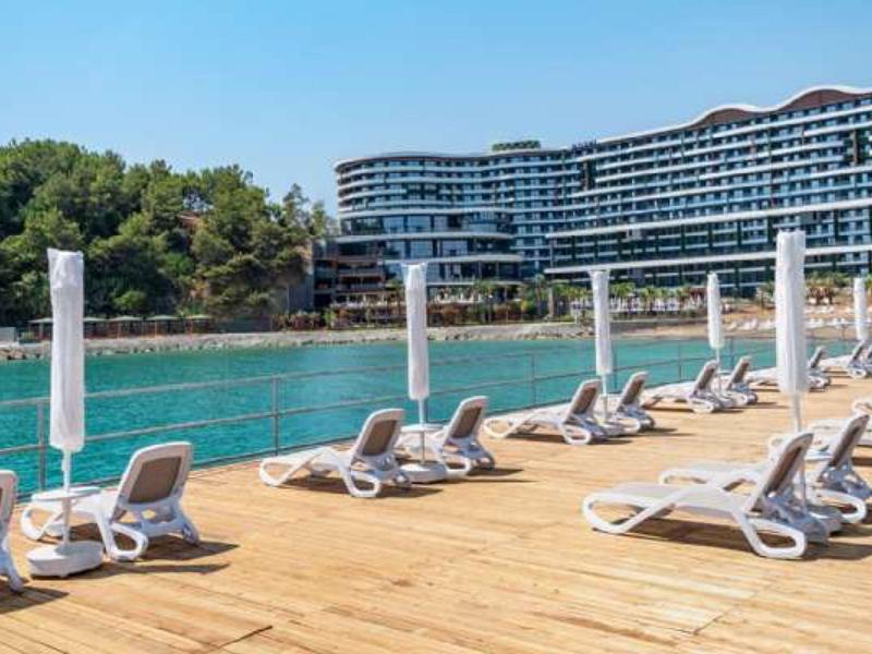 Sonnige Holzterrasse mit Liegestühlen am klaren Wasser vor modernem Hotel und grünen Bäumen.