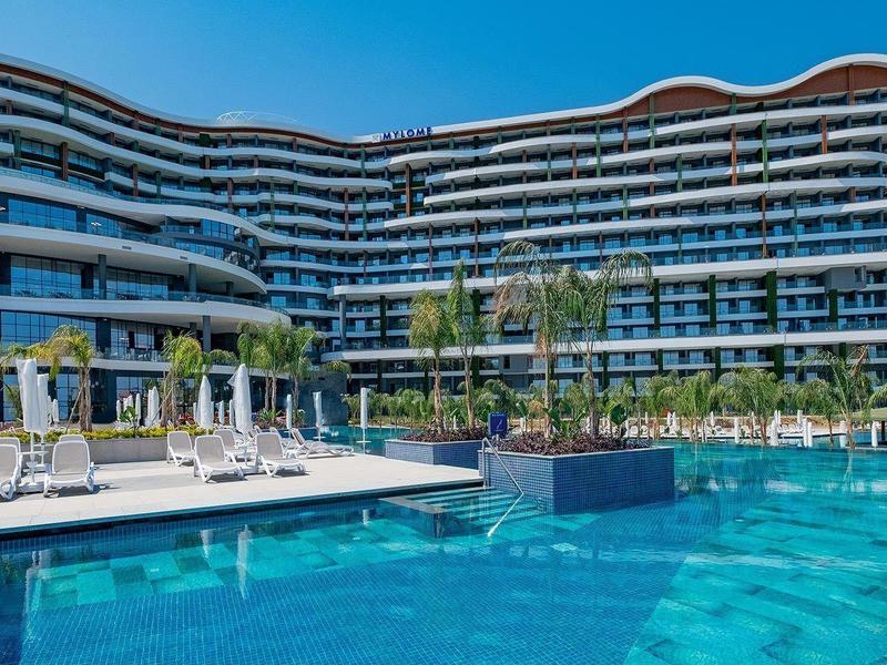 Modernes Hotel mit geschwungenem Design, großem Pool, Liegestühlen und Palmen unter blauem Himmel.