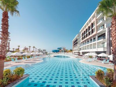 Grande piscina all'aperto con palme e lettini davanti a un moderno edificio hotel con cielo sereno.