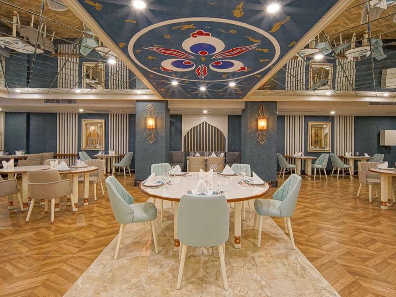 Elegancka jadalnia z okrągłymi stołami, jasnoniebieskimi krzesłami i ozdobnym sufitem.