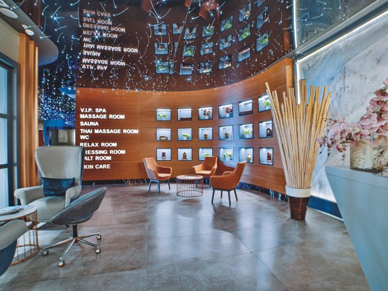 Nowoczesny salon hotelowy z stylowymi fotelami, dużą dekoracją roślinną i cyfrową ścianą informacyjną.