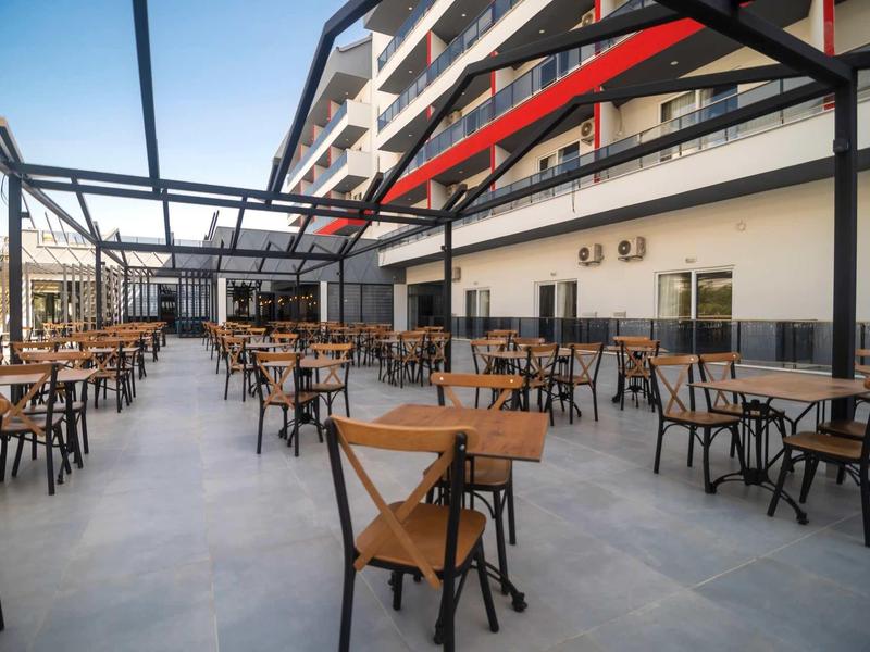 Gran terraza con mesas y sillas de madera frente a un edificio de hotel con balcones.