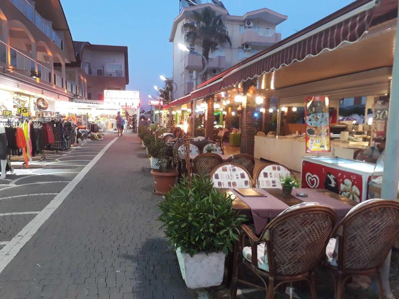 Straße mit Geschäften und Außenbewirtung, beleuchtet, mit Pflanzen und bunter Dekoration am Abend.