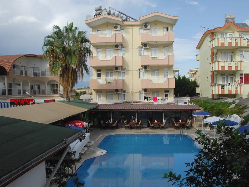 Vierstöckiges Hotel mit Balkonen, Pool im Vordergrund, Palmen und blauer Himmel.