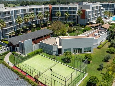 Modern hotelcomplex met tennisbaan, zwembad en groene omgeving onder een blauwe lucht.
