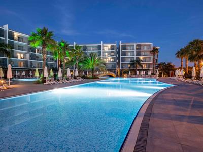 Moderner Außenpool bei Nacht mit Beleuchtung, Palmen und Loungemöbeln vor einem Hotelgebäude.