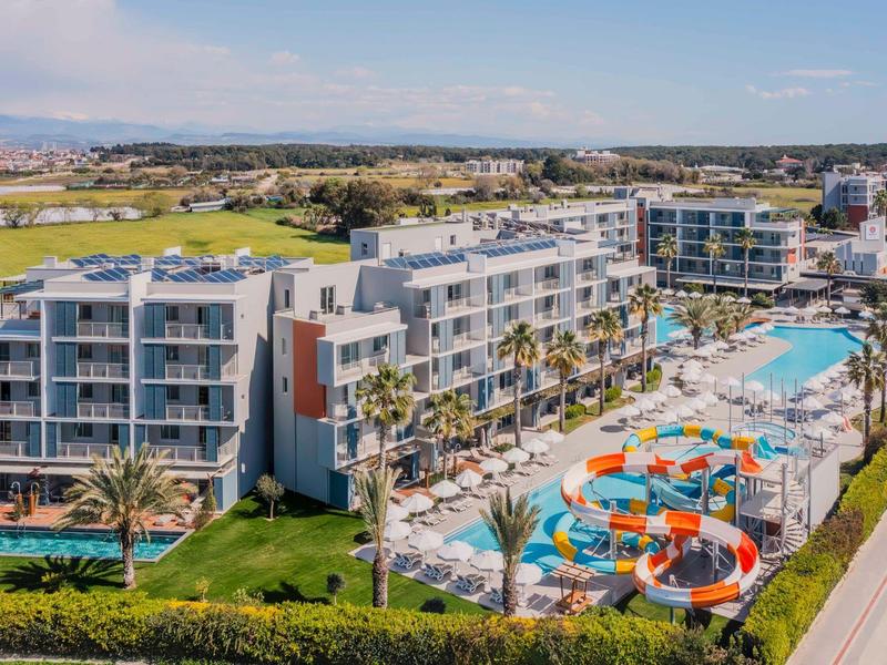 Moderní hotelový komplex s bazénem, vodními skluzavkami a okolní zelení pod modrou oblohou.