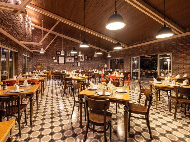 Příjemná restaurace s dřevěnými stoly a židlemi, osvětlená závěsnými lampami v místnosti s vysokými stropy a cihlovými stěnami.