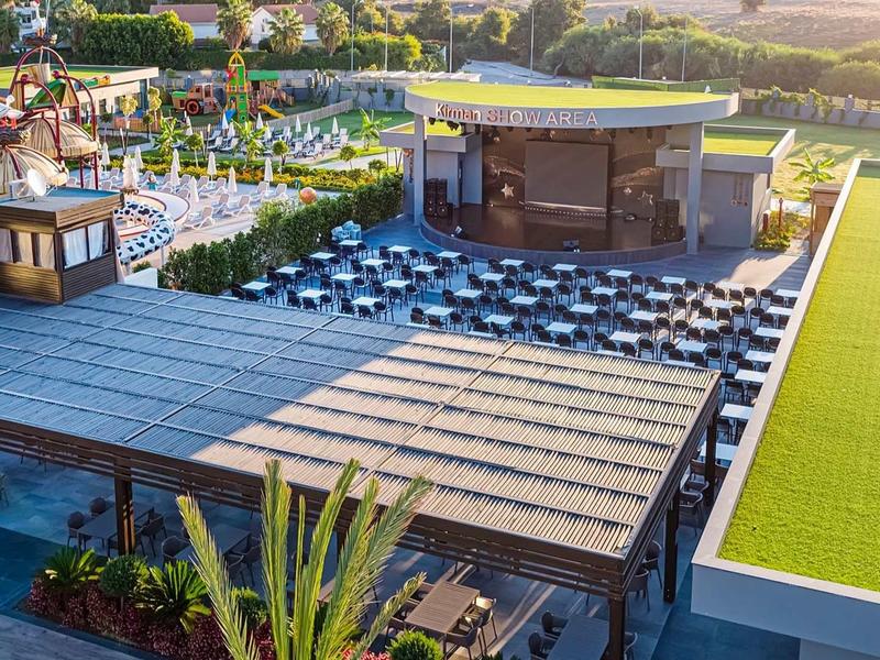 Große Terrasse mit Tischen, Stühlen und Sonnenschirmen neben grünem Feld und Gebäuden bei Sonnenschein.