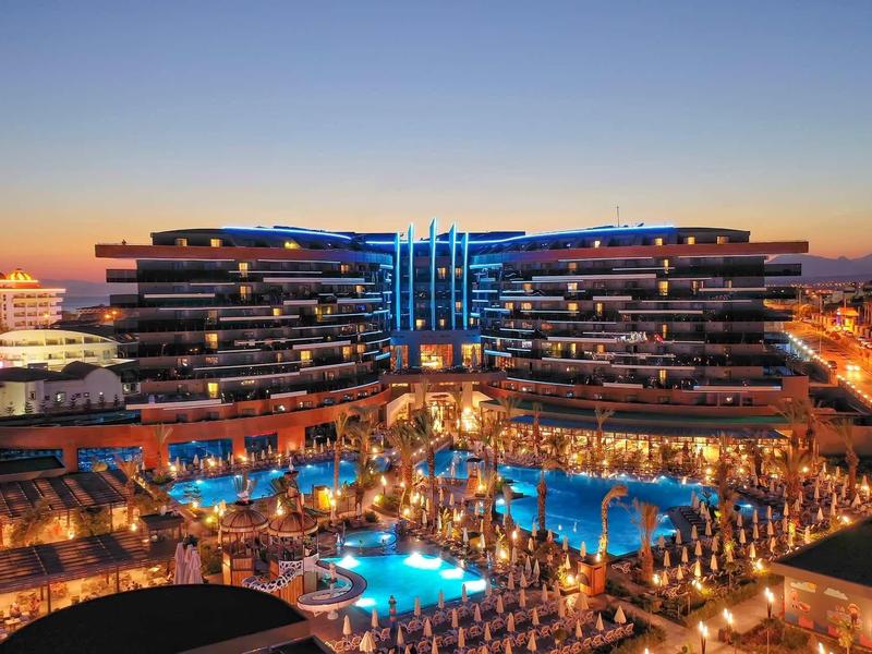 Großes Hotel mit blau beleuchtetem Pool und modernen, terrassenförmigen Gebäuden bei Sonnenuntergang.