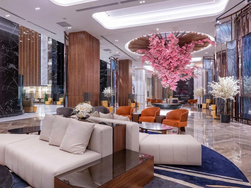 Moderner Hotel-Lobby mit weißen Sofas, orangenen Sesseln, Holzsäulen und großer rosa Blumeninstallation.