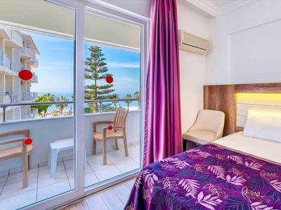 Helles Hotelzimmer mit Balkon, violettem Bettüberwurf, beigen Stühlen und Meerblick.
