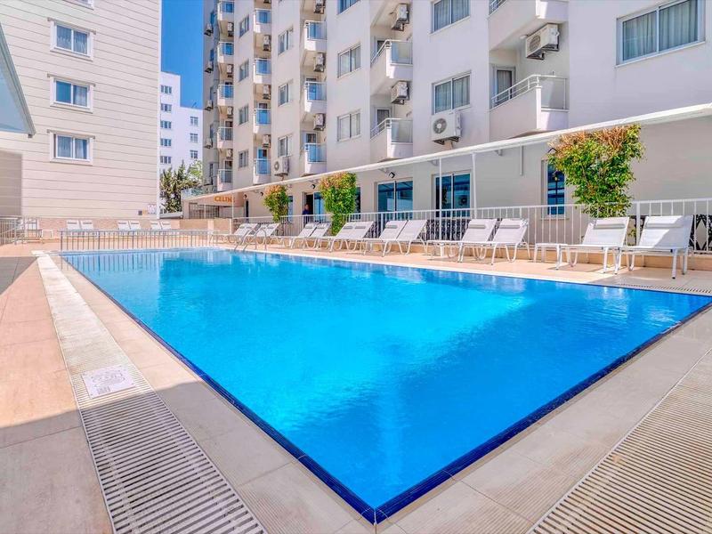 Piscina all'aperto moderna davanti a un hotel pluripiano con lettini e ombrelloni.