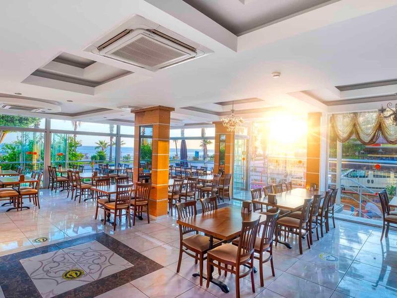 Area colazione luminosa con sedie e tavoli in legno, grandi finestre e sole.