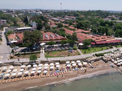 Plaża z parasolami i leżakami przed czerwonymi budynkami hotelowymi w zielonym otoczeniu.