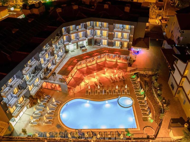 Nocne zdjęcie z lotu ptaka oświetlonego basenu hotelowego otoczonego leżakami i budynkami.