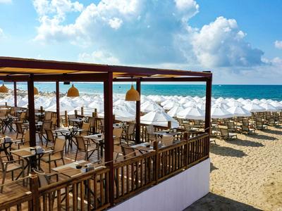 Restaurant de plage avec terrasse en bois à côté d'une plage de sable avec parasols blancs sous ciel bleu.