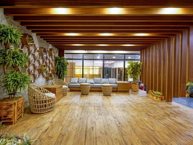 Hotellounge met houten vloer, plafondbalken, planten en rotan zitmeubels bij grote ramen.