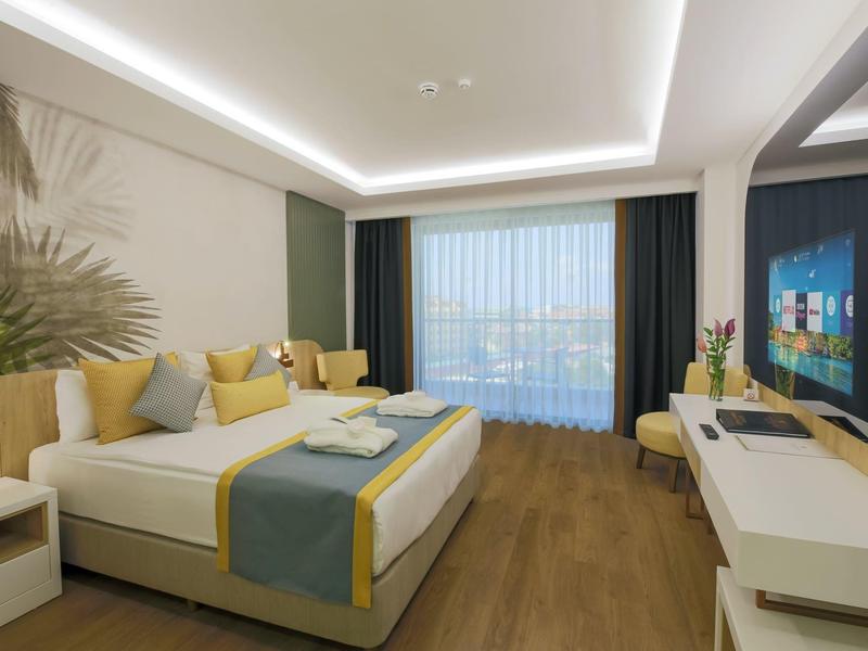 Een moderne hotelkamer met een groot bed, houten vloer en een raam met doorschijnende gordijnen.