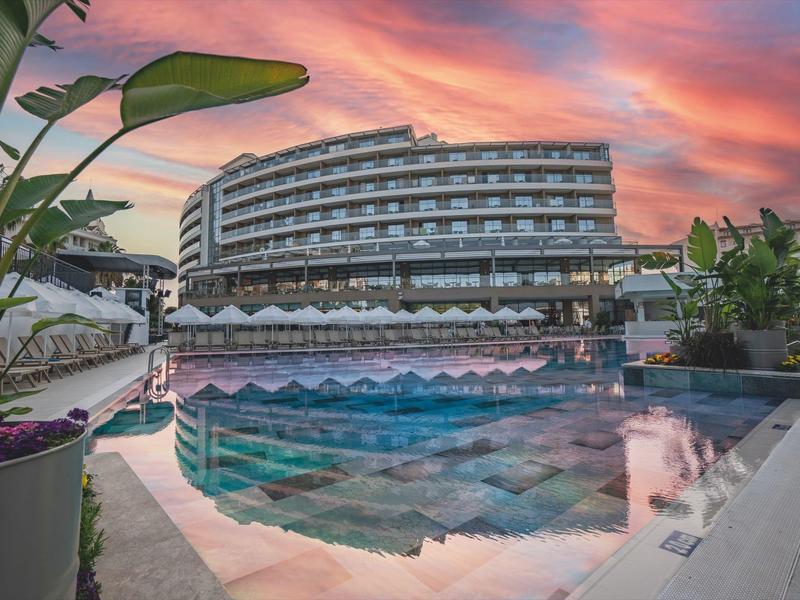 Gevormd hotelgebouw bij zonsondergang met zwembad dat kleurrijke lucht en planten weerspiegelt.
