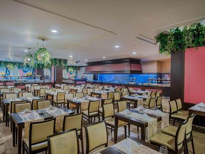 Velká, dobře osvětlená jídelna s mnoha stoly a židlemi, zdobená rostlinami.