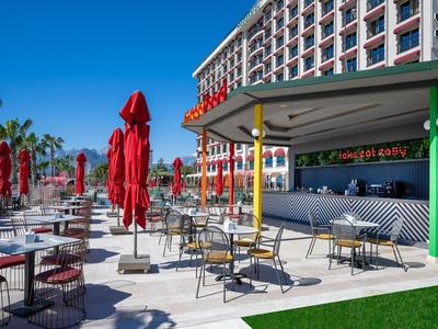 Area esterna di un hotel con tavoli, sedie e ombrelloni rossi sotto un cielo blu.
