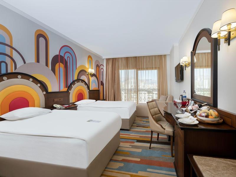 Moderní hotelový pokoj se dvěma postelemi, psacím stolem, židlemi a barevným vzorem na zdi.