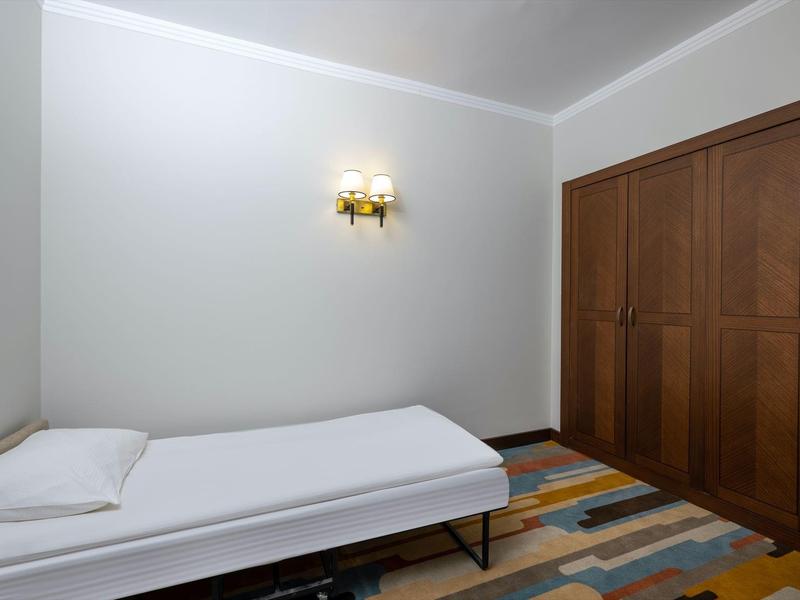 Jednoduchý hotelový pokoj s jednolůžkem, barevným kobercem a velkou dřevěnou skříní.