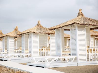 Weiße Strandkabinen mit Strohdächern und Liegen stehen nebeneinander am Kiesstrand.