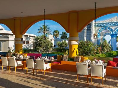 Area lounge aperta con sedute colorate sotto archi con vista su architettura e palme.