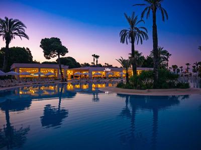 Hotel serale con piscina illuminata e palme sotto un cielo limpido.