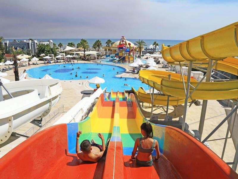 Twee kinderen op kleurrijke glijbanen met uitzicht op een zwembad en zee bij een vakantieoord.