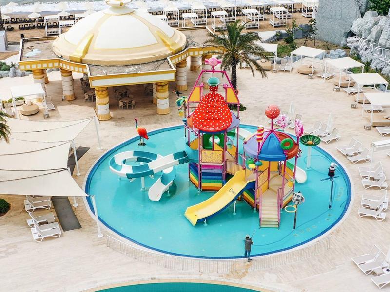 Kleurrijke kinderspeelplaats met glijbanen en waterpartijen naast zwembad en ligstoelen.