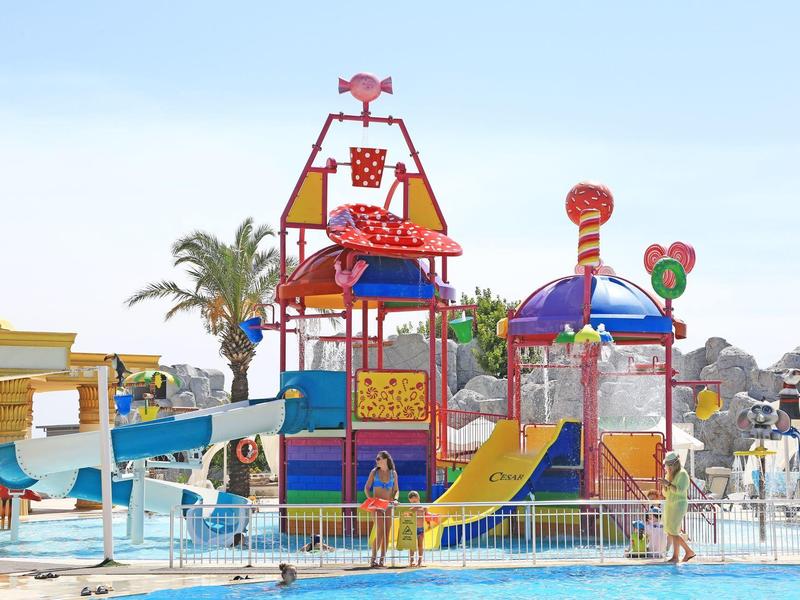 Kleurrijke waterspeelstructuur met glijbanen en fonteinen bij hotelzwembad onder blauwe lucht.